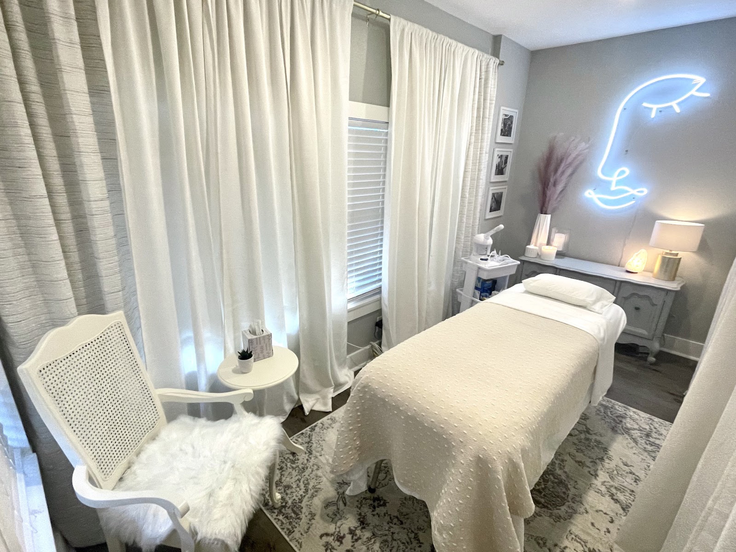 The treatment room: a made-up facial bed, soft white curtains and a glowing line-drawn face sign on the wall.
